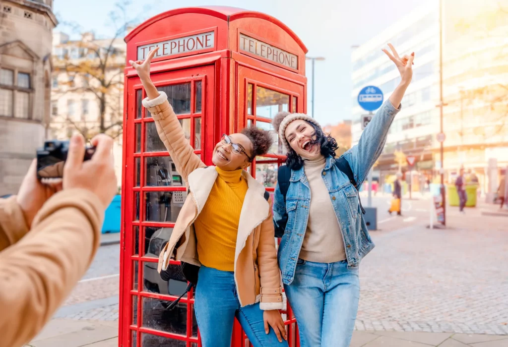 Two girls stood in front of a red telephone box and smiling at a camera